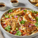 A plate of vibrant Dumpling Salad with crispy golden-brown dumplings placed on top of a colorful mix of cabbage, carrots, and fresh cilantro, garnished with sesame seeds and served with dipping sauce.